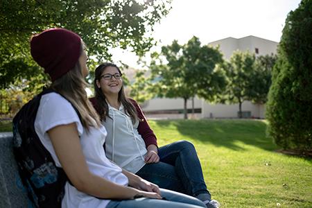 Image of two female students sitting on a bench and talking. In the image background the silhouette of a building can be seen.